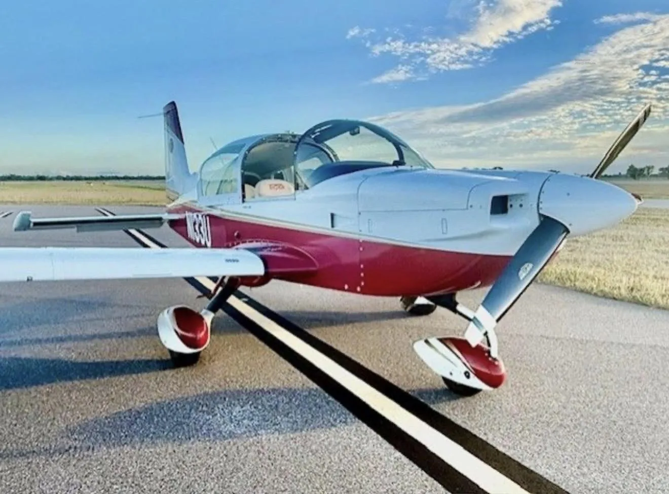 A red and white single-engine aircraft with a bubble canopy is parked on an asphalt runway under a blue sky.
