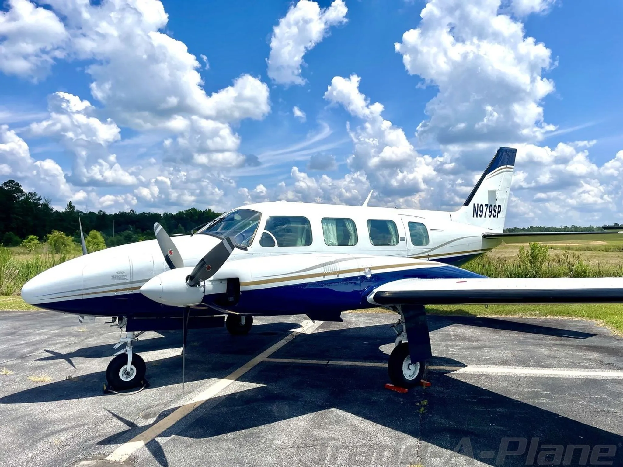 A white and blue twin-engine Piper aircraft, N979SP, is parked on an asphalt ramp under a blue sky with white clouds.