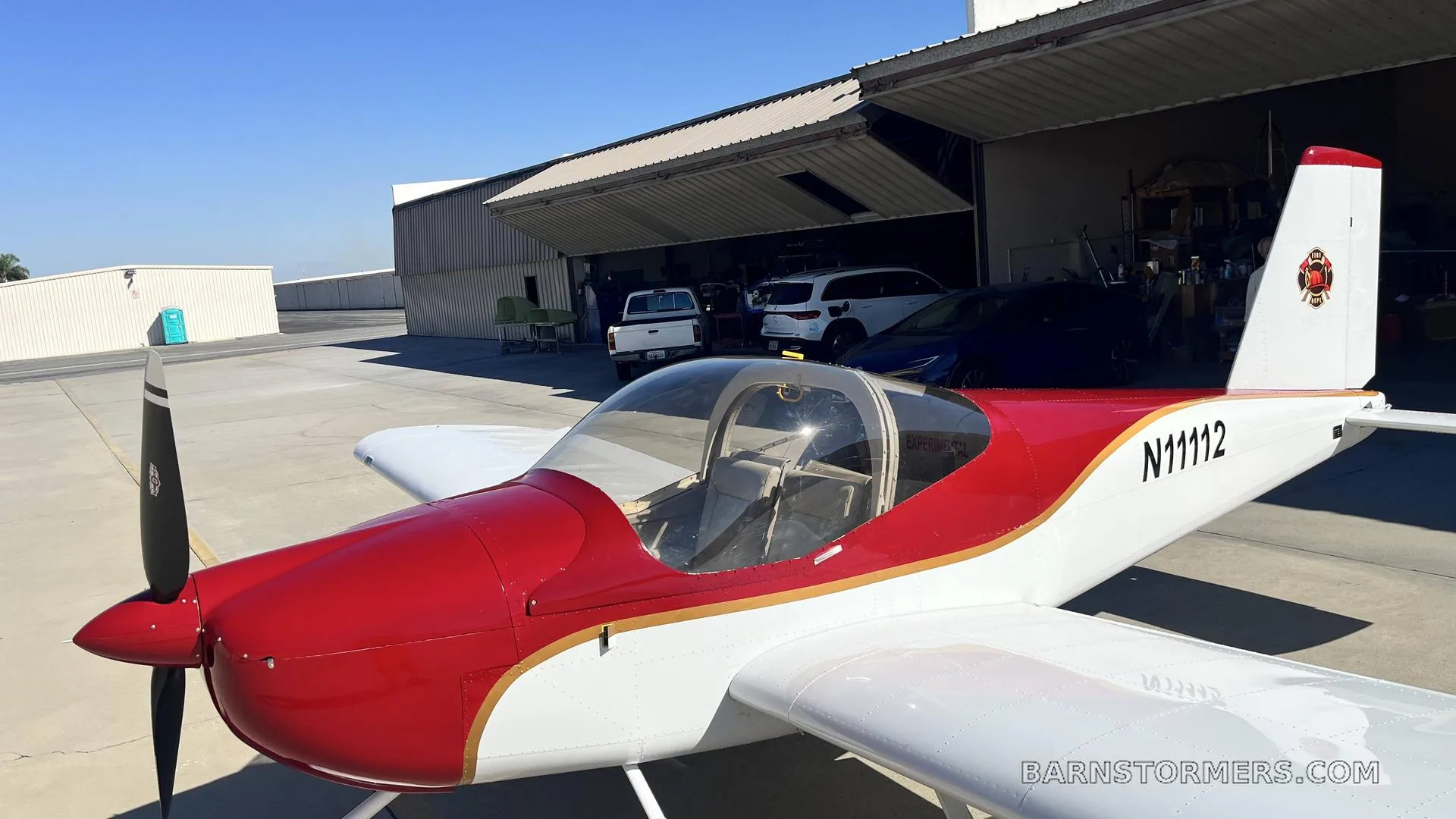 A red and white single-engine aircraft with registration N11112 is parked on an airfield apron in front of an open hangar.
