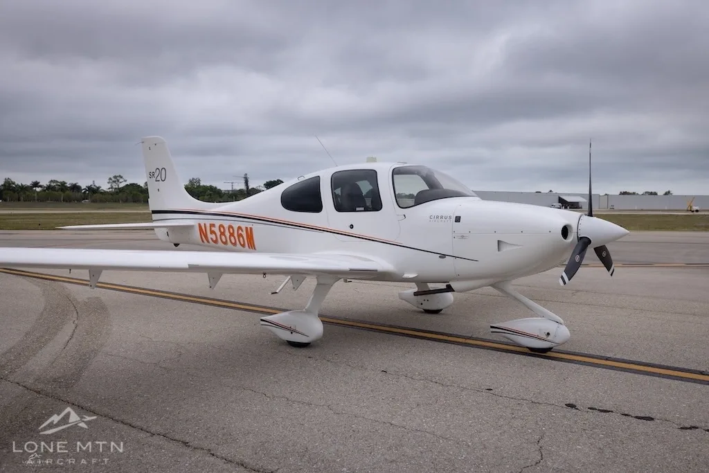 A front-side view of a white Cirrus SR20 aircraft with orange and black stripes, registration N5886M, parked on an airfield.