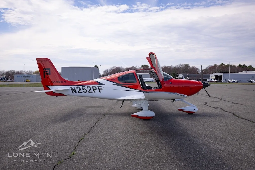 A side view of a red and white Cirrus SR20 aircraft with its doors open, parked on an airfield.