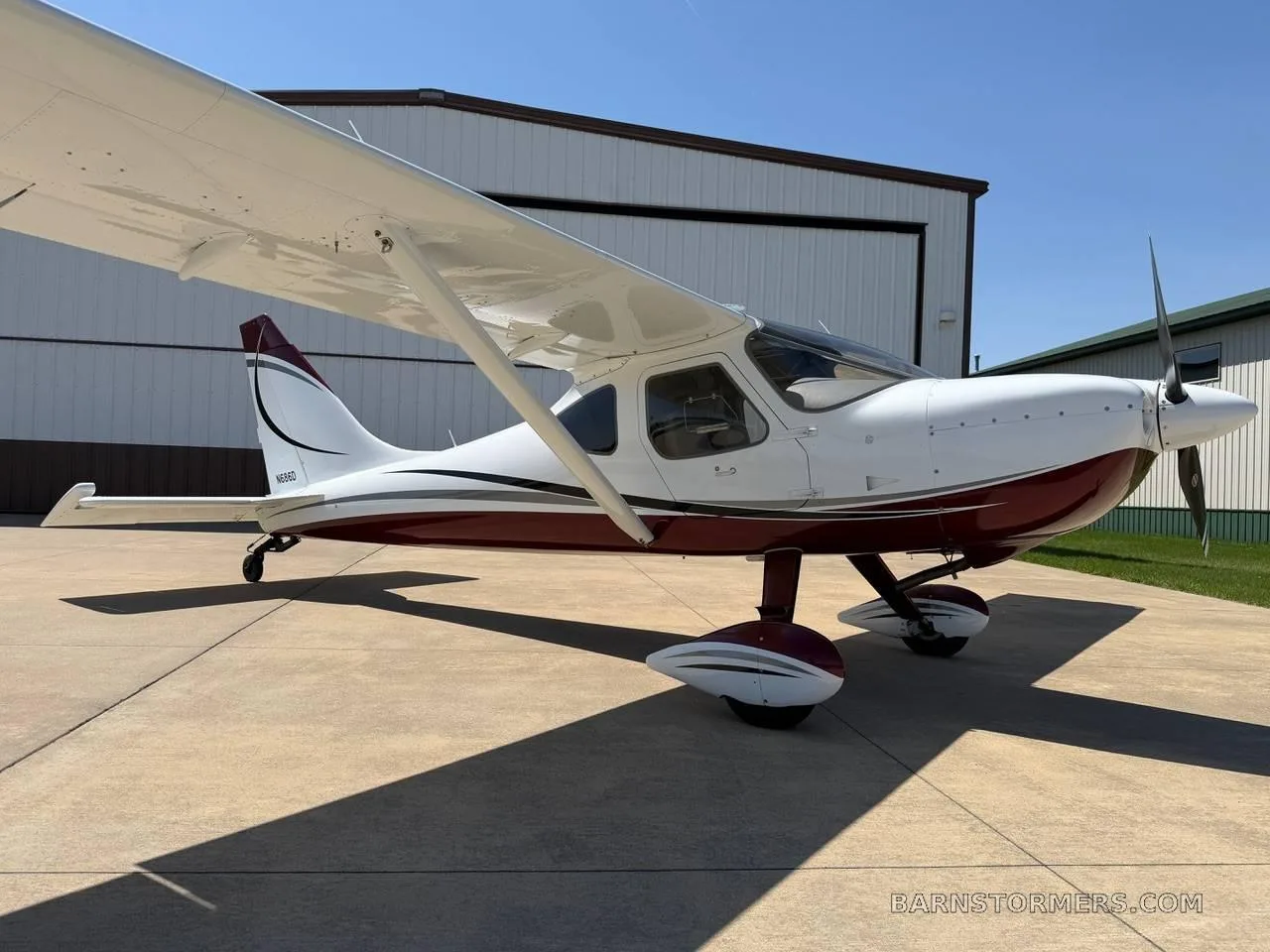 A side view of a white and maroon high-wing single-engine aircraft (N686D) parked on a concrete tarmac next to a hangar.