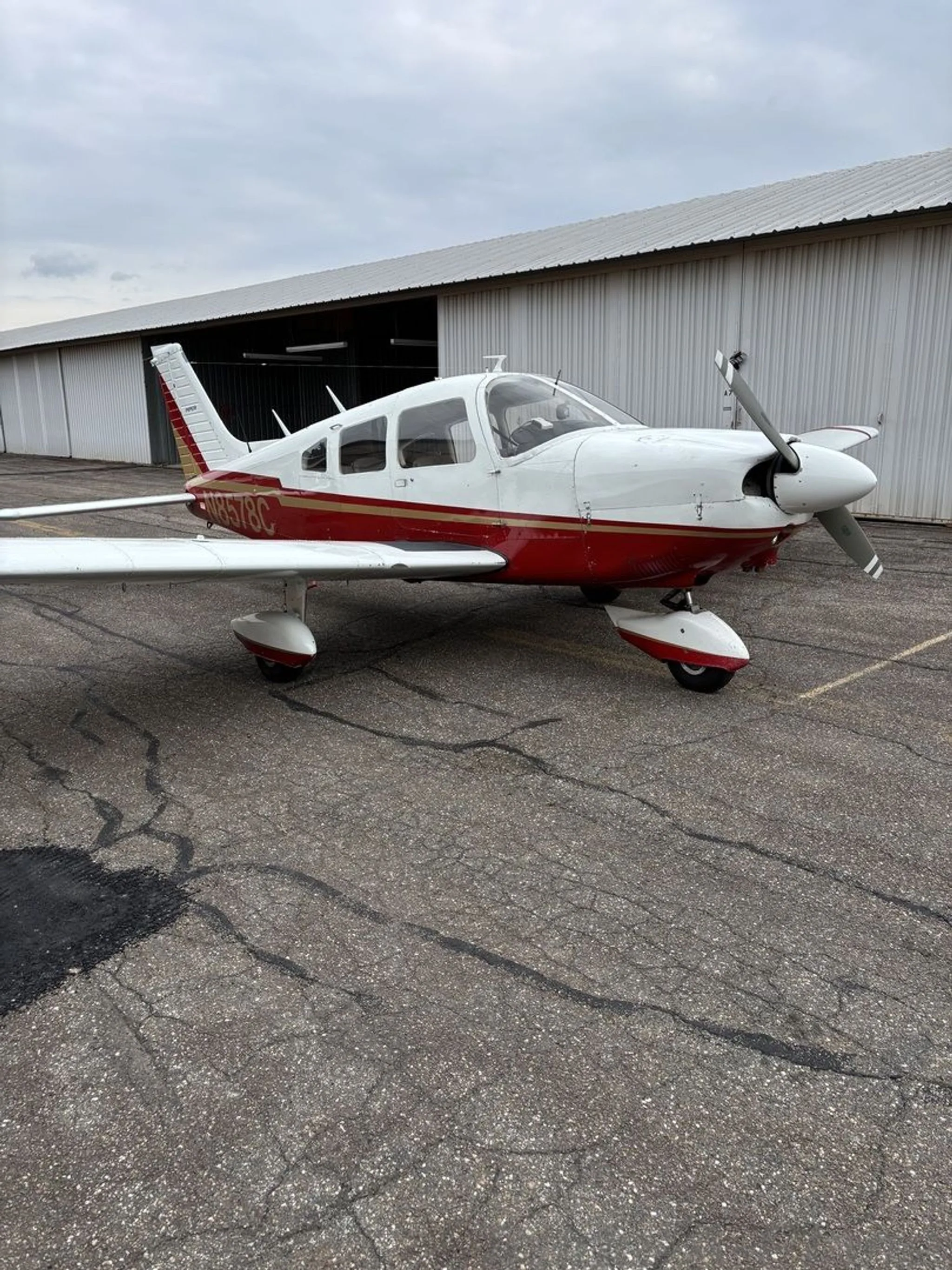 A white Piper aircraft with red and gold livery, N8578C, parked on an asphalt surface in front of a hangar.