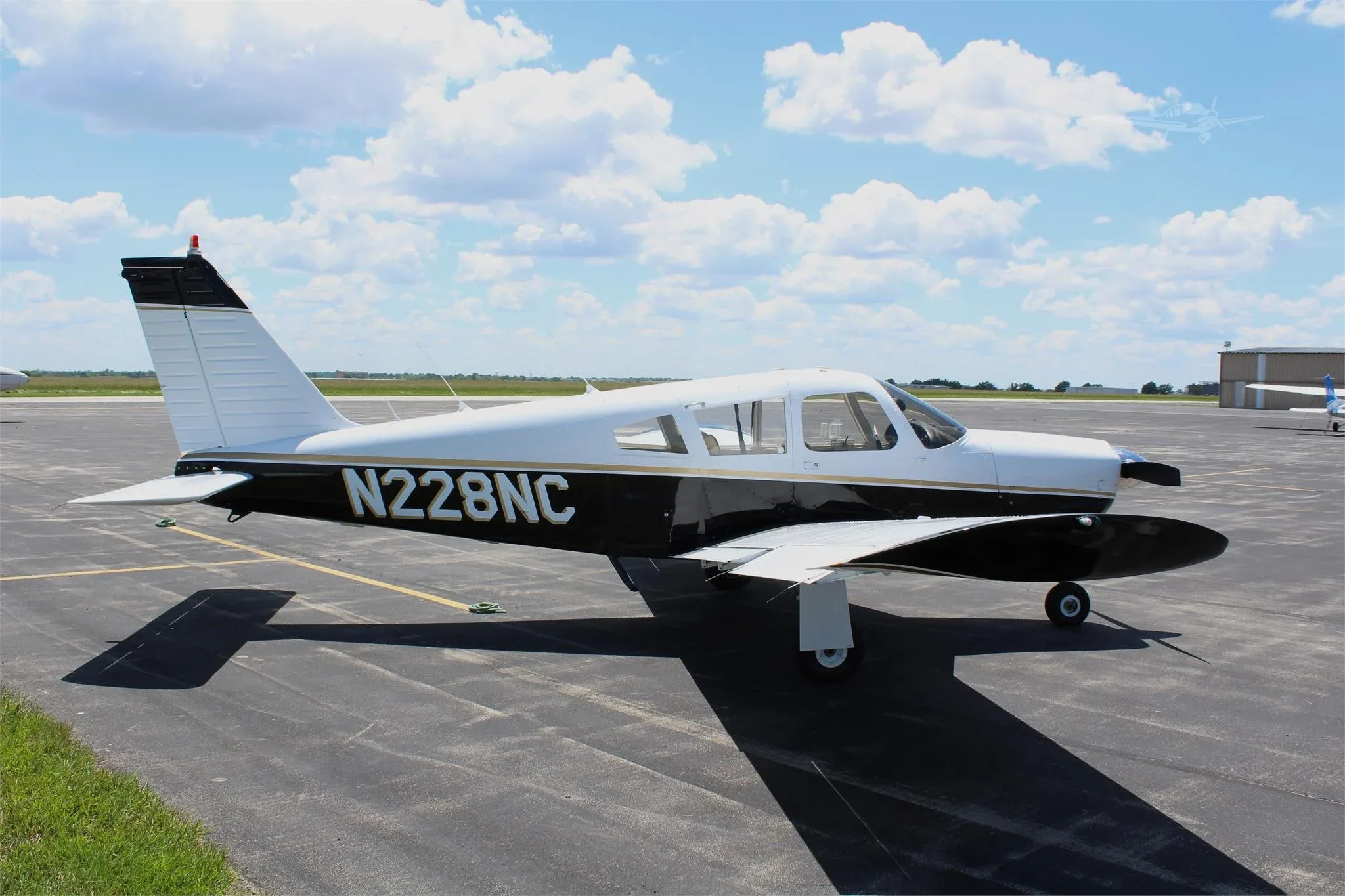 A white and black Piper PA-28 aircraft, registration N228NC, parked on an airport tarmac under a partly cloudy sky.