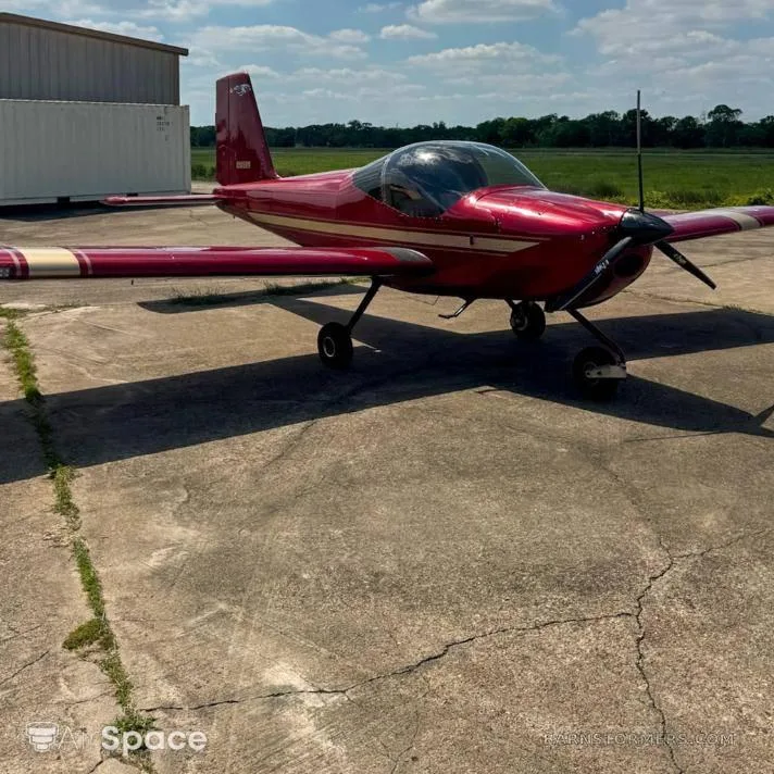 A red Vans RV-6 aircraft with white stripes parked on a concrete tarmac next to a hangar under a partly cloudy sky.