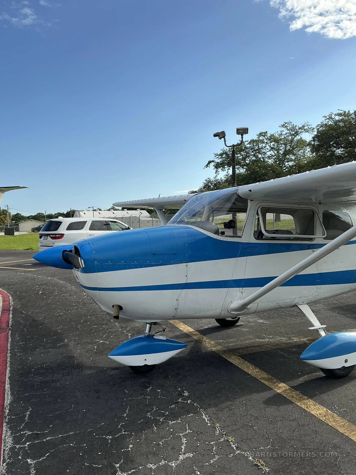 A white and blue Cessna 172 aircraft is parked on an asphalt ramp, viewed from the front-right.