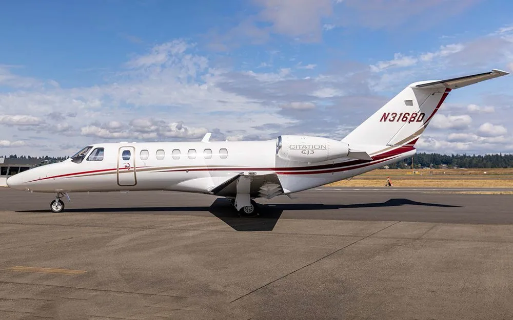 A white Cessna Citation CJ3 business jet with red and gold stripes on the tarmac under a partly cloudy sky.
