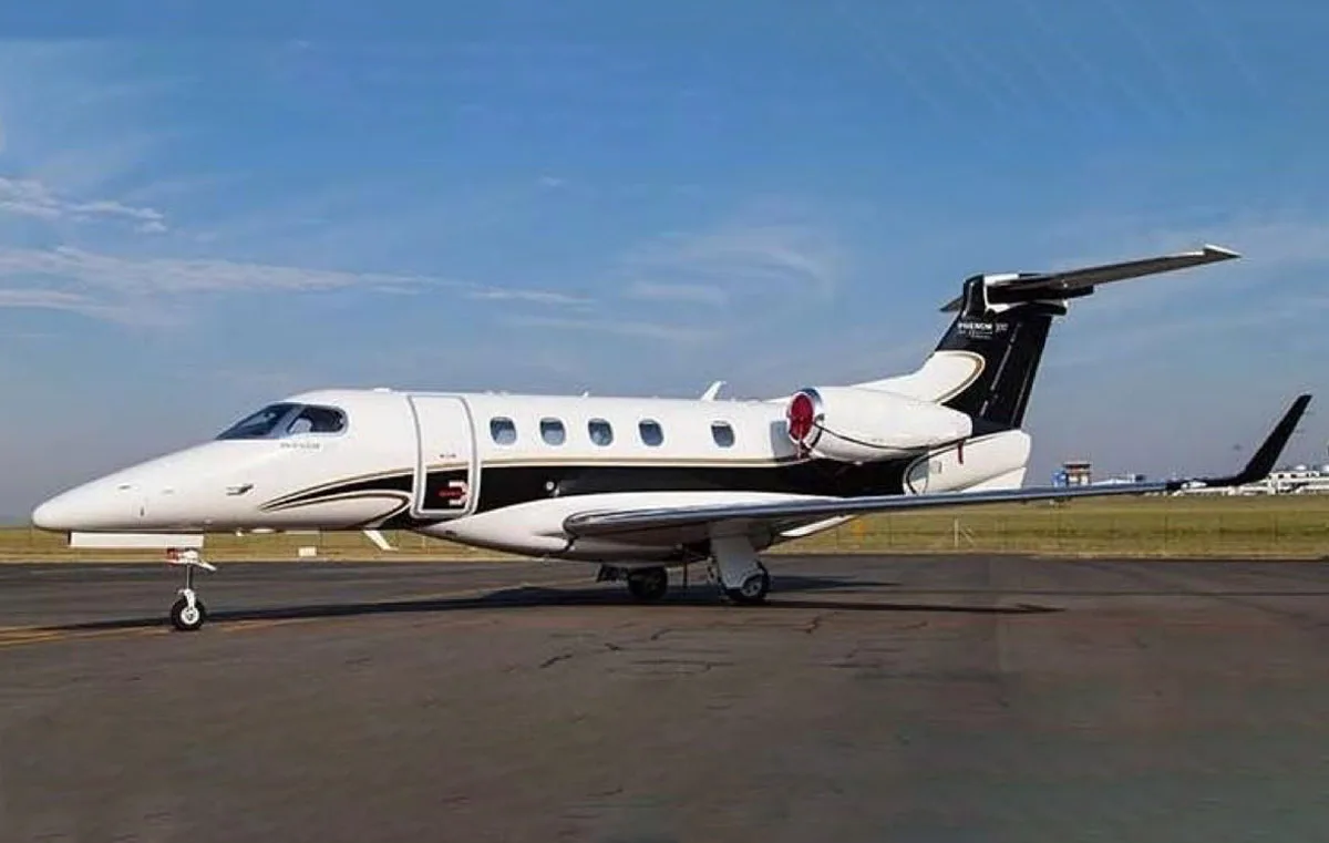 A white Embraer Phenom 300 business jet with black and gold stripes is parked on an airport tarmac under a blue sky.