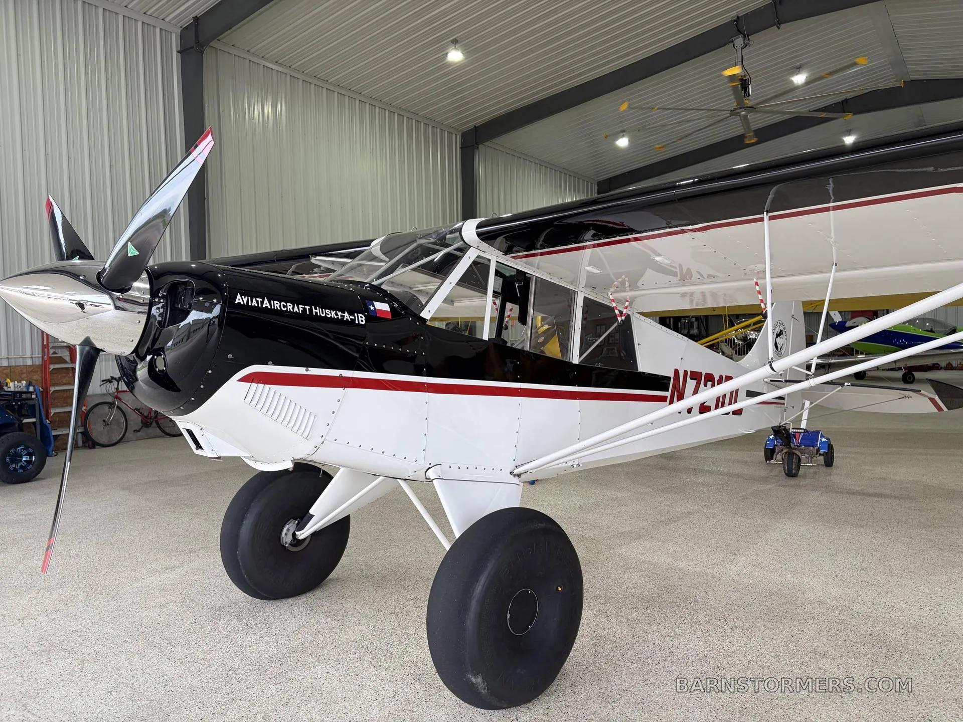 A close-up view of the front right side of a black and white Aviat Aircraft Husky A-1B with red stripes and large tundra tires, inside a hangar.