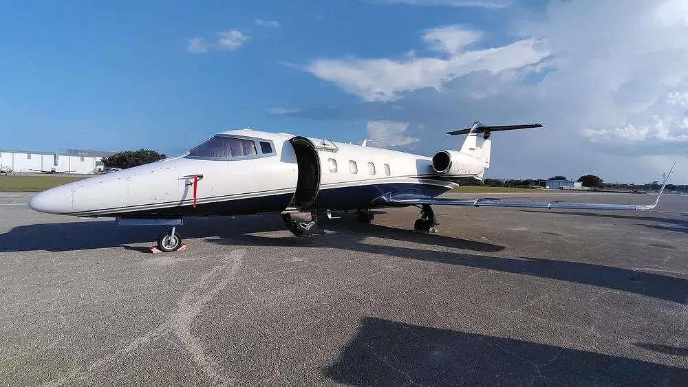 A white and dark blue private jet with its main door open, parked on an asphalt tarmac under a partly cloudy sky.