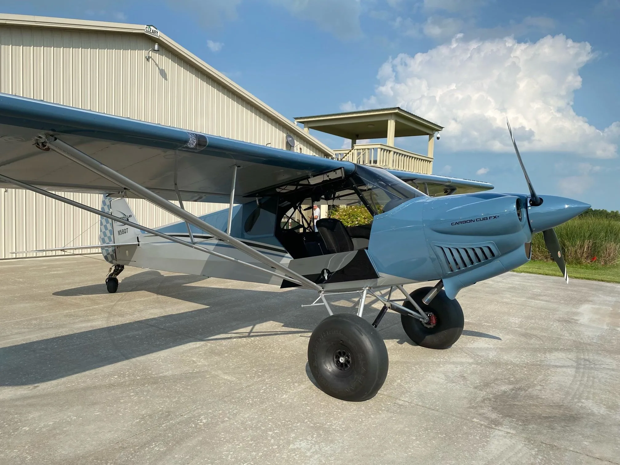 A blue and grey Carbon Cub FX-3 aircraft with registration N58QT parked outside a hangar under a partly cloudy sky.