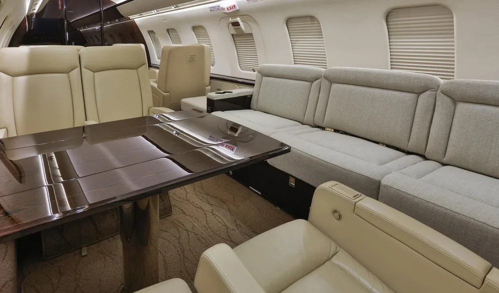 A close-up view of the private jet's cabin, highlighting the dark wood table and the light grey sofa seating area.