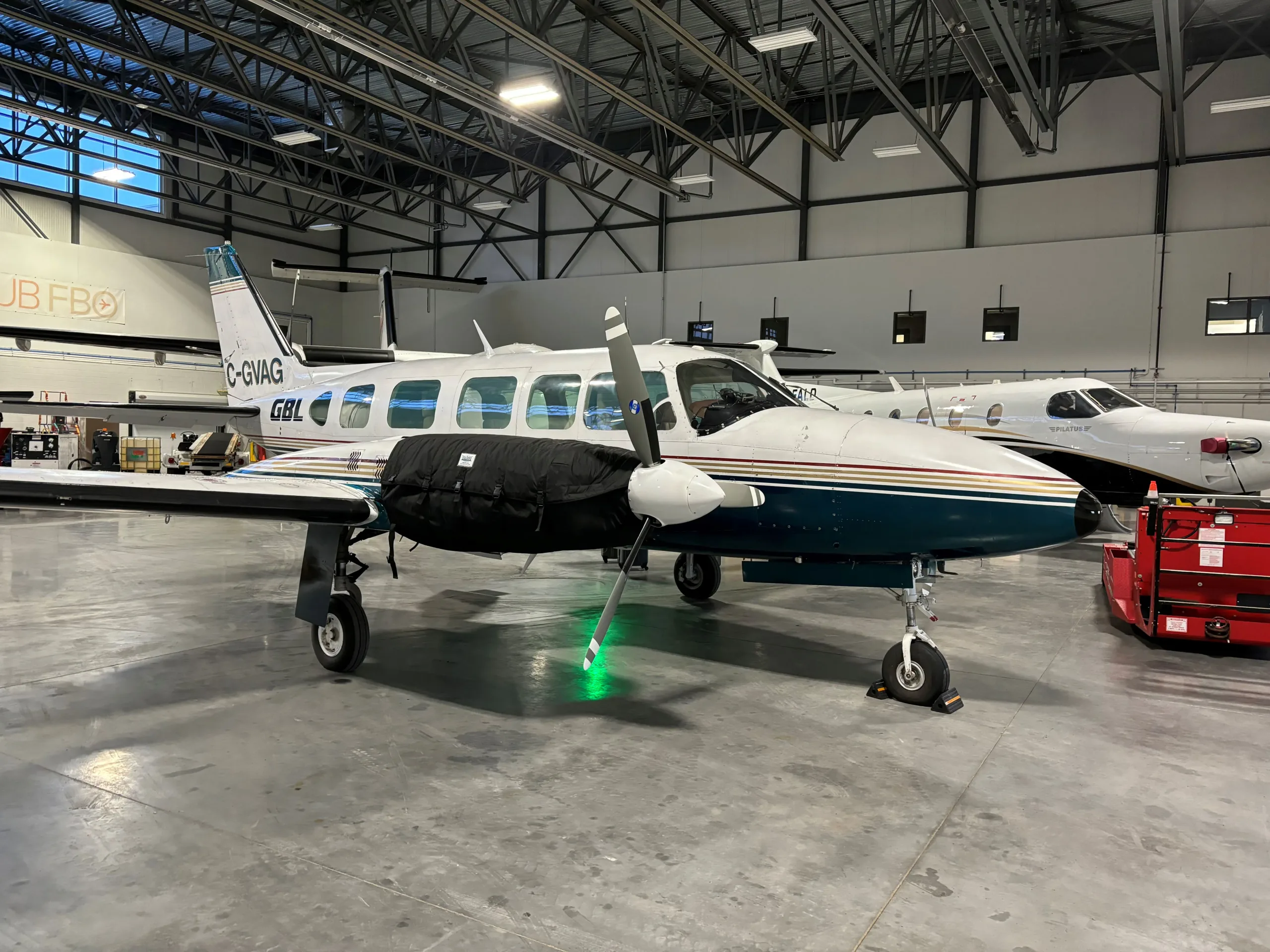 A white twin-engine propeller aircraft with dark green and gold stripes, registration C-GVAG, parked in a hangar with engine covers on.