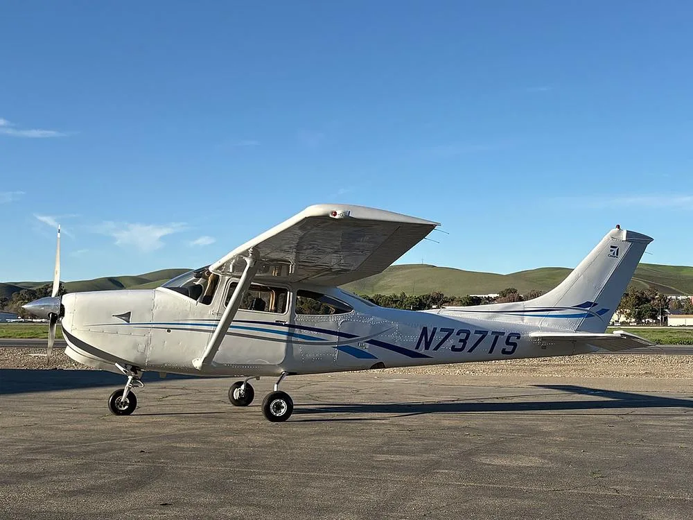A white Cessna 182 aircraft with blue stripes, registration N737TS, parked on an airfield with rolling hills in the background.
