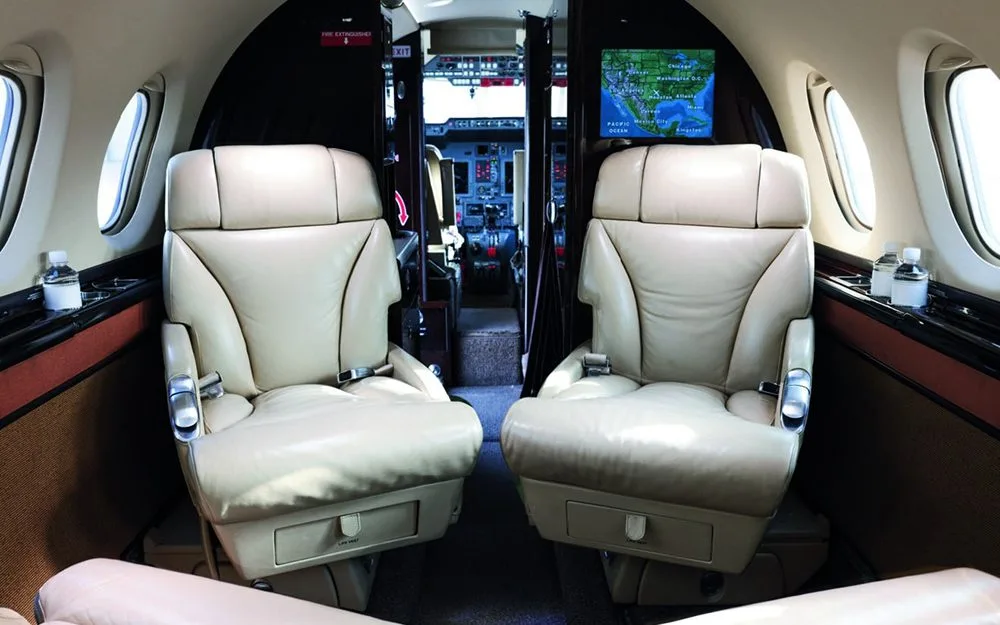 The aircraft cabin features two cream-colored leather seats facing forward, with the cockpit visible through an open doorway.