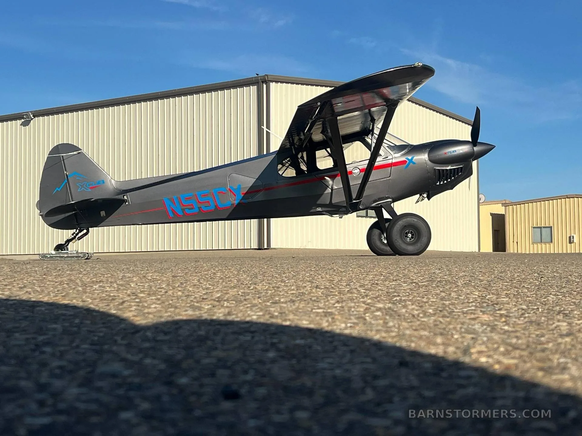 A low-angle side view of a dark gray X-Cub aircraft with red and blue accents, registration N55CX, parked on an asphalt ramp.