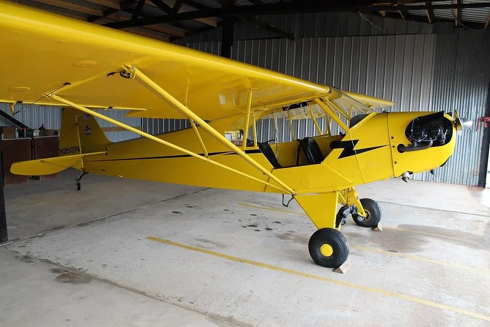 A yellow Piper Cub-style aircraft with a black lightning bolt stripe is parked in a hangar, viewed from the front-right side.