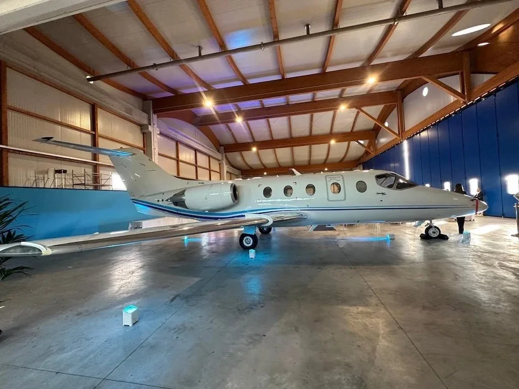 A white business jet with blue stripes is parked inside a hangar with its landing gear illuminated by blue lights.