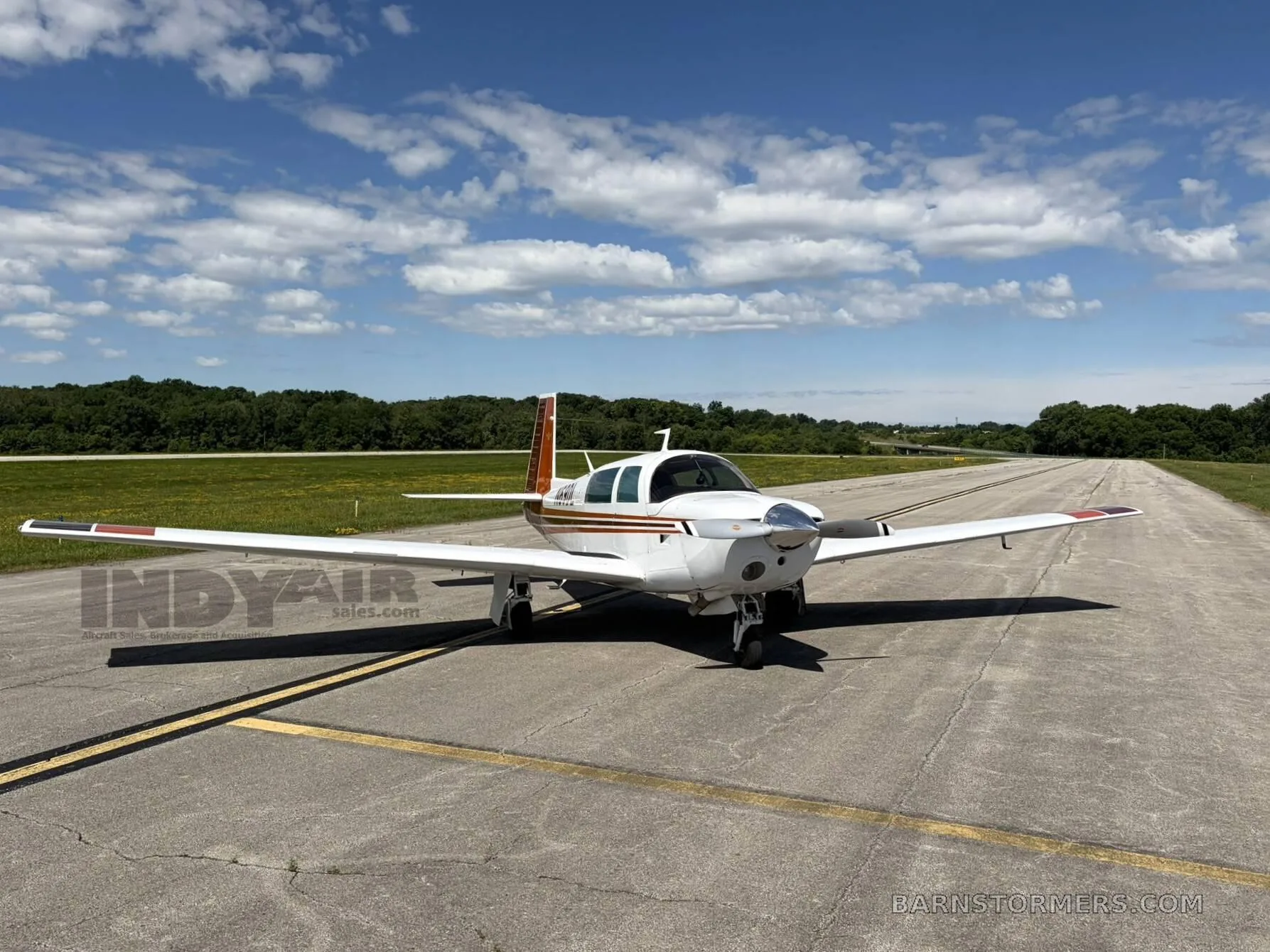 A white and brown Mooney M20J aircraft is parked on an asphalt runway under a partly cloudy sky, viewed from the front.