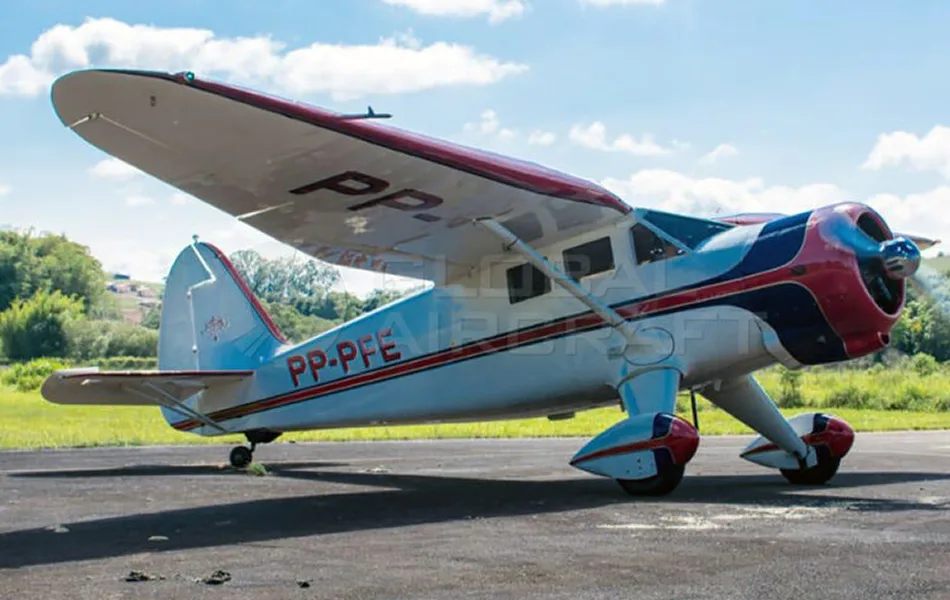 A white, red, and blue vintage high-wing monoplane with registration PP-PFE parked on an airfield under a blue sky.
