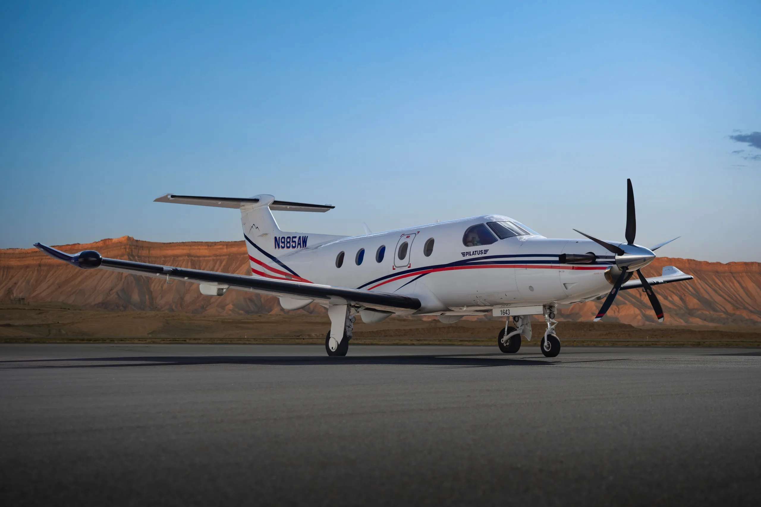 A Pilatus PC-12 aircraft, N985AW, with a white, blue, and red paint scheme, sits on a tarmac with mountains in the background under a clear sky.