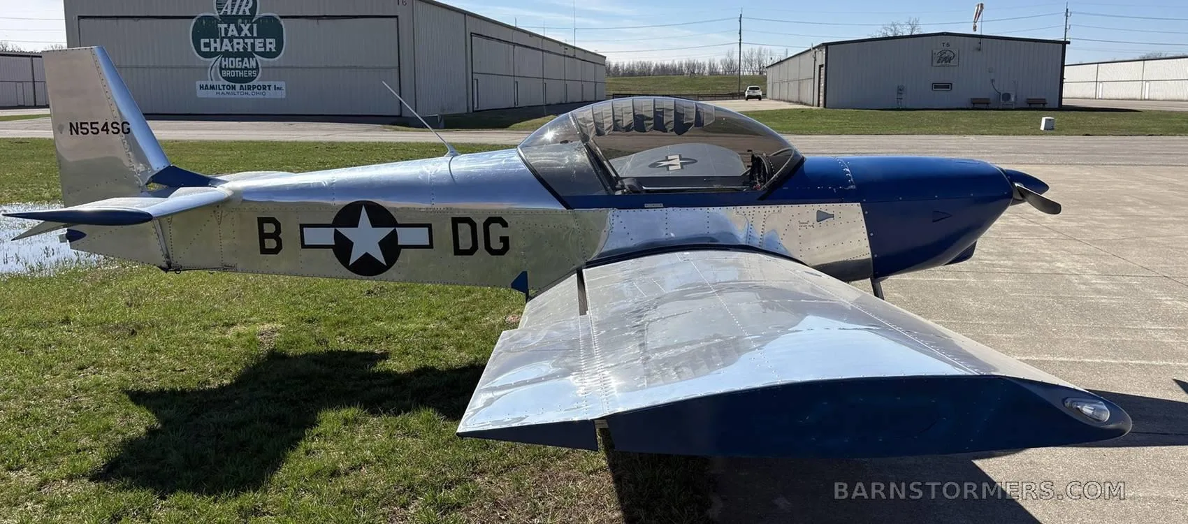 A blue and polished aluminum single-engine aircraft with a bubble canopy and "B * DG" markings on the side, parked on an airfield.
