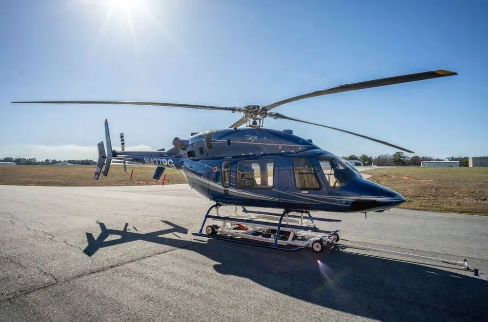 A blue Bell 427 helicopter with white and silver stripes is parked on an asphalt runway under a bright sun.