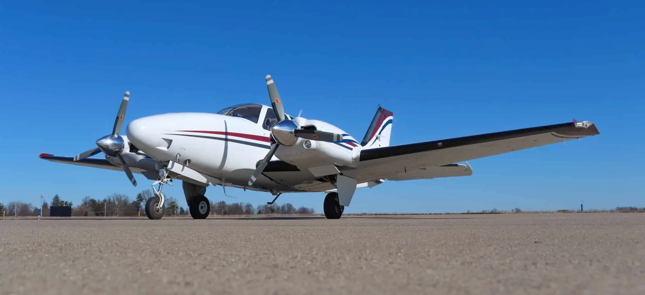 Low angle front-side view of a white twin-engine Beechcraft Baron N204G with red, blue, and gray stripes, parked on an airfield.