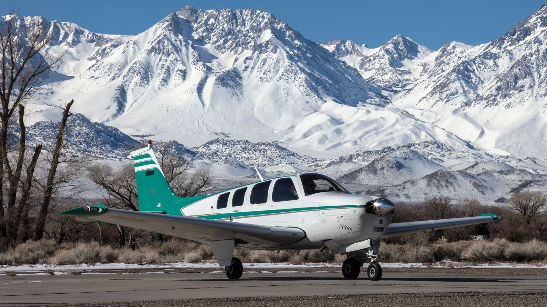 A white and teal Beechcraft Bonanza sits on a runway with snow-covered mountains in the background.
