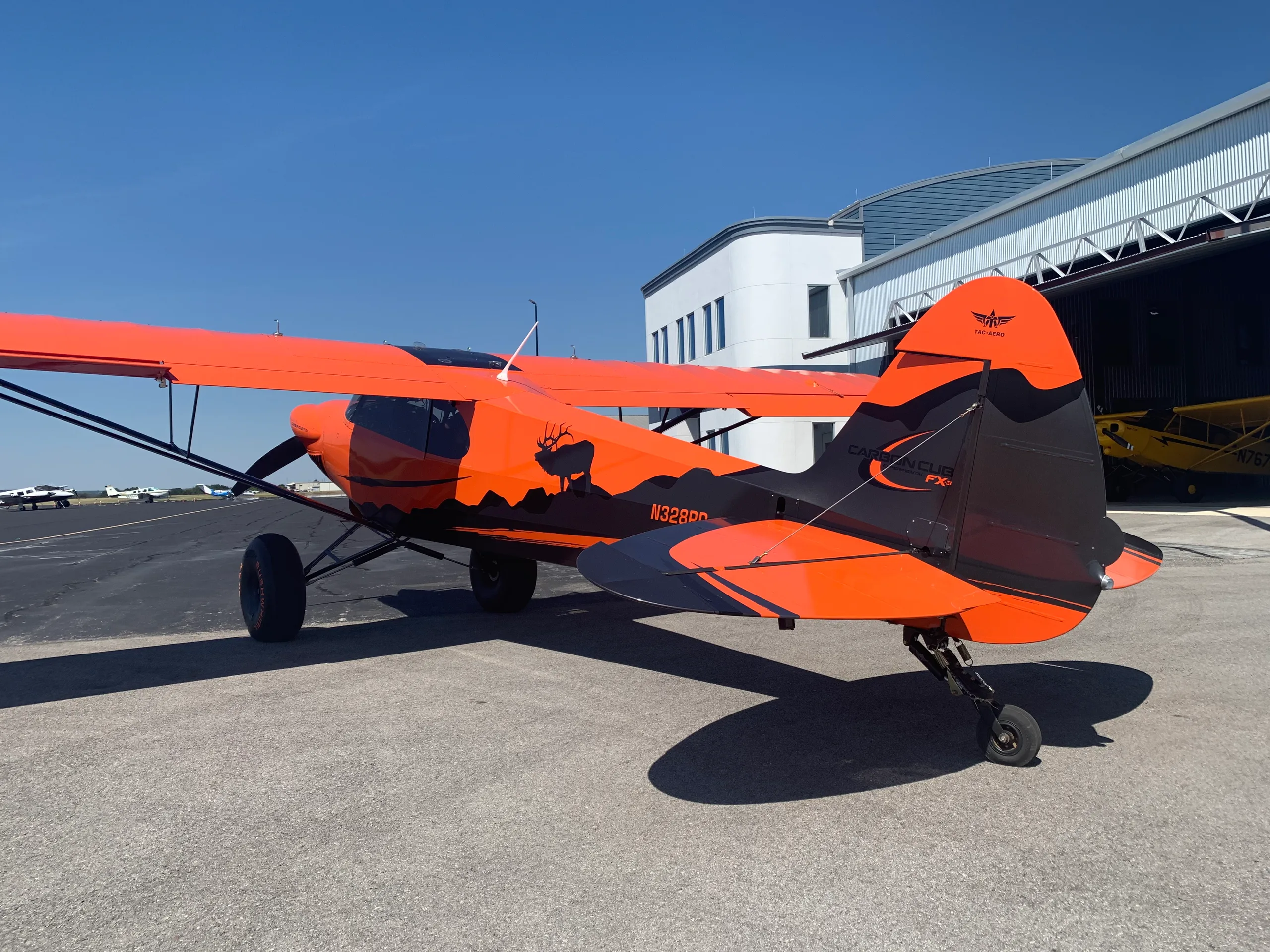 An orange and black Carbon Cub FX-3 aircraft with an elk silhouette on the tail, parked on an airport tarmac next to a hangar.