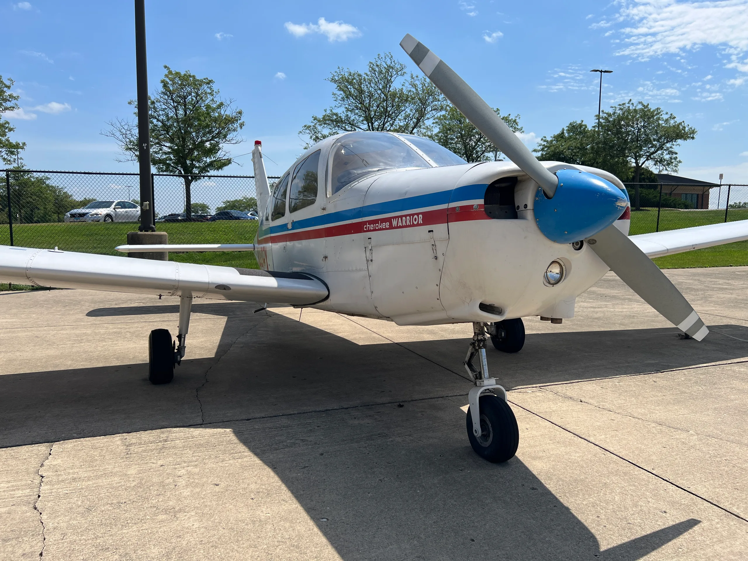 A white Piper Cherokee Warrior aircraft with blue and red stripes is parked on a concrete surface under a clear sky, viewed from the front-right.