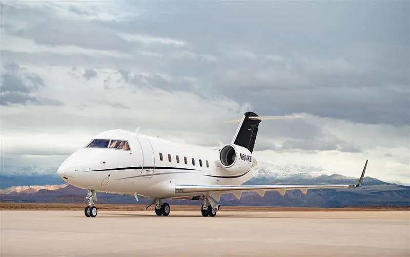 A white Bombardier Challenger 604 business jet with black accents, tail number N604KE, parked on an airfield with mountains in the background.