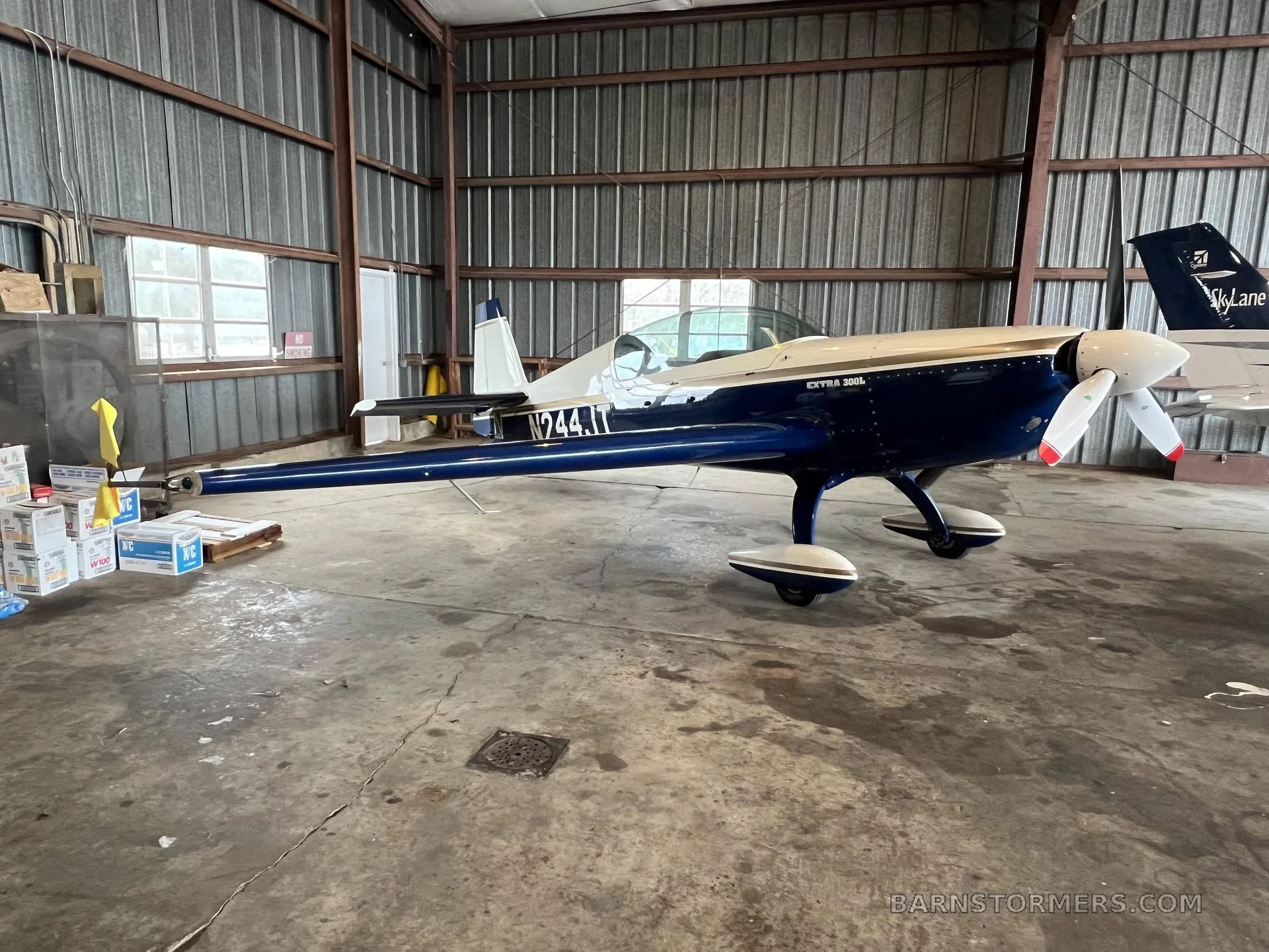 A blue and white Extra 300L aircraft with tail number N244JT is parked inside a metal hangar, viewed from the front-right.