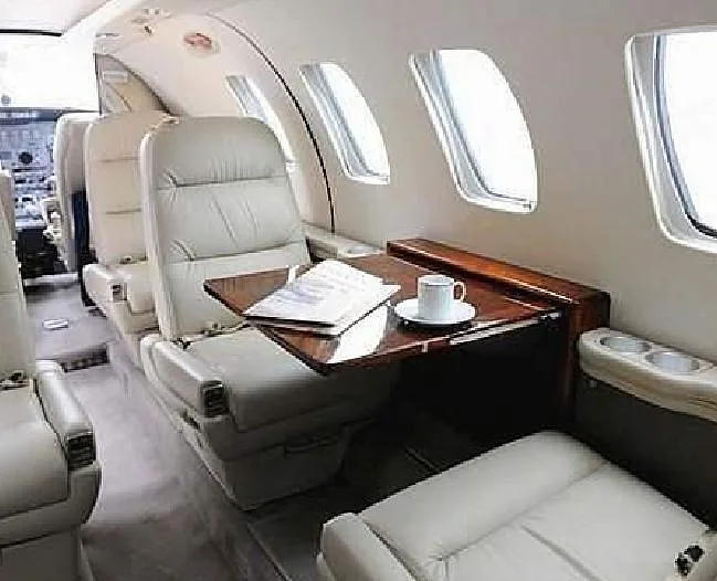 A view of the aircraft cabin interior showing light-colored leather seats, a wooden table with a cup and papers, and windows.
