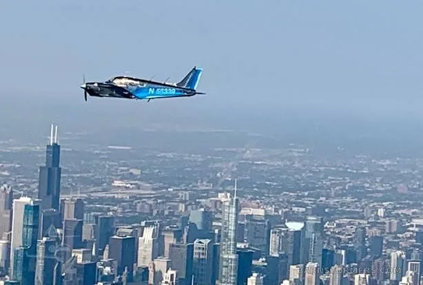 A dark blue single-engine propeller aircraft with registration N 56339 flies over a city skyline on a clear day.