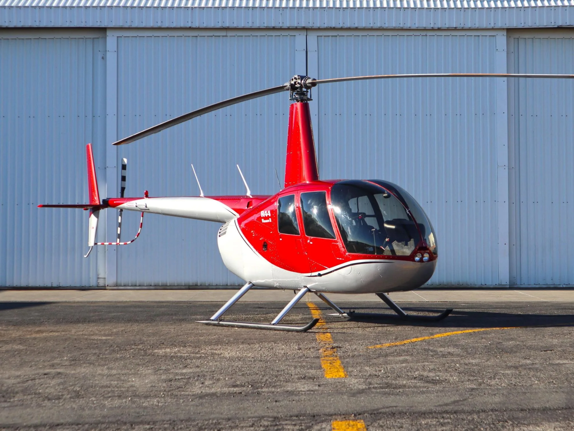 A red and white Robinson R44 helicopter is parked on an asphalt tarmac in front of a white hangar.