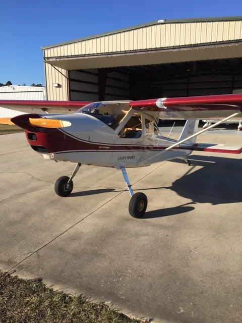 A white and red light sport aircraft is parked on a concrete tarmac in front of an open hangar under a clear blue sky.