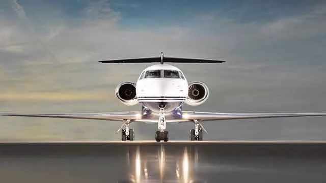 A white business jet with dark engines and tail, viewed head-on on a reflective tarmac under a dramatic sky.