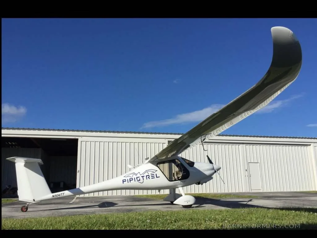 A white Pipistrel aircraft with its wing folded up, parked on grass next to a white hangar under a blue sky.