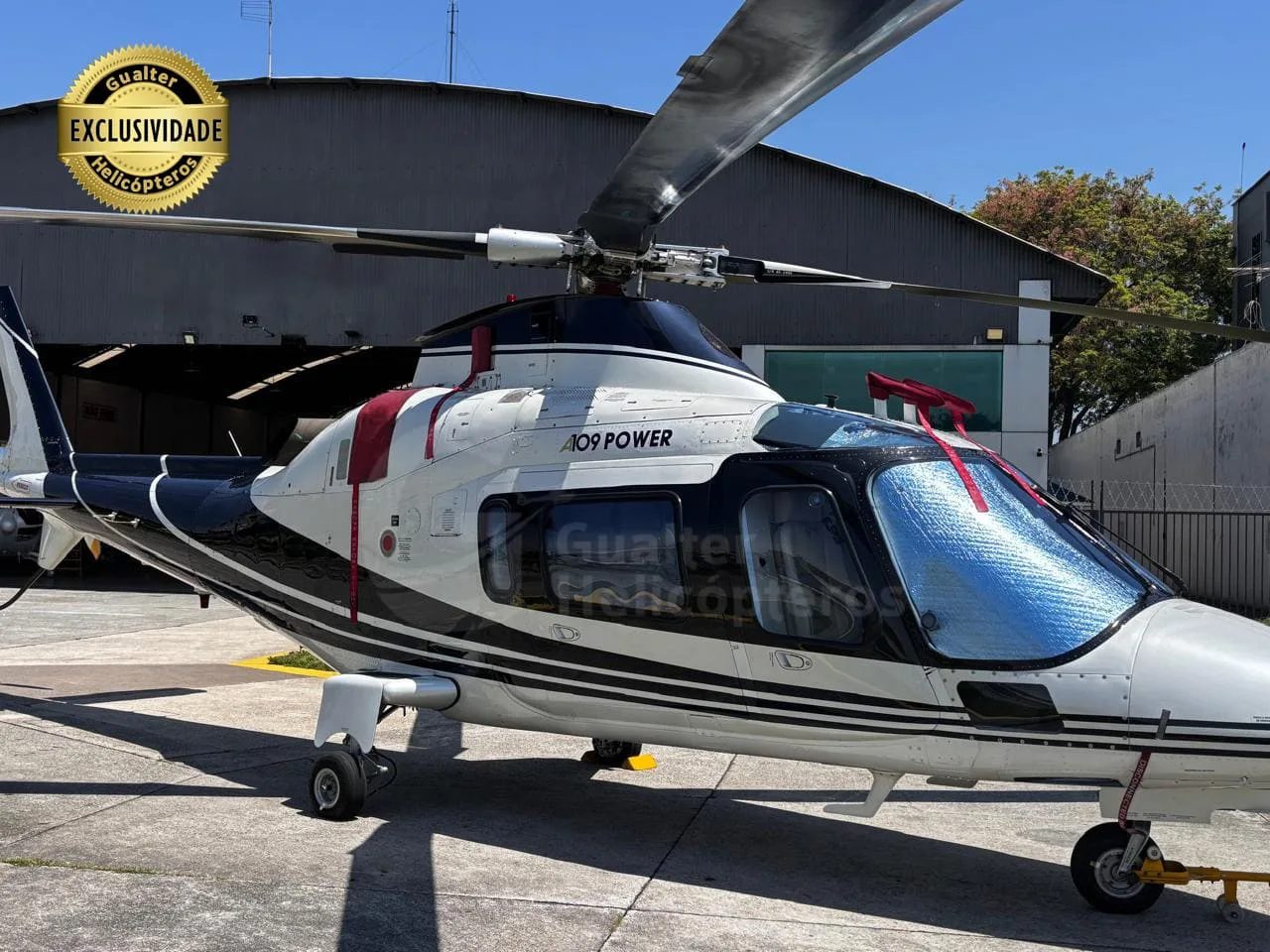 A white and black Agusta A109 Power helicopter with a sunshade on the windshield, parked on an asphalt apron.