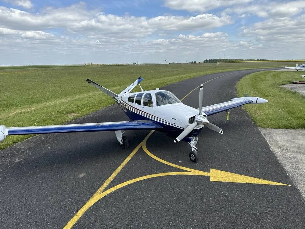 A front-quarter view of a white and blue Beechcraft Bonanza aircraft on an airfield, with green fields and a cloudy sky.