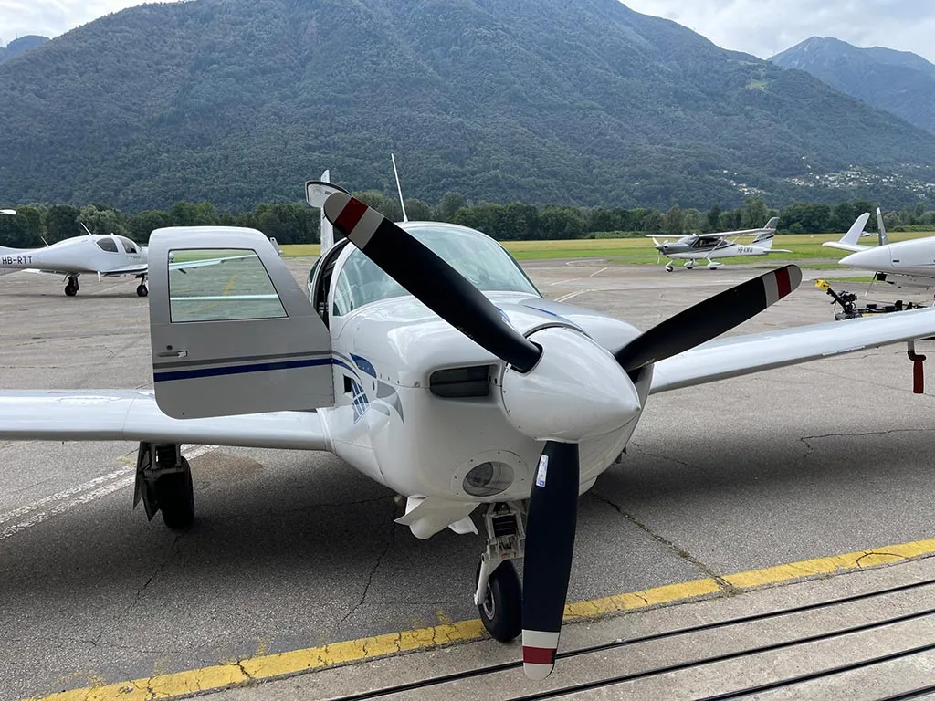 Front view of a white single-engine propeller aircraft with blue stripes, parked on an airfield with its door open and mountains in the background.