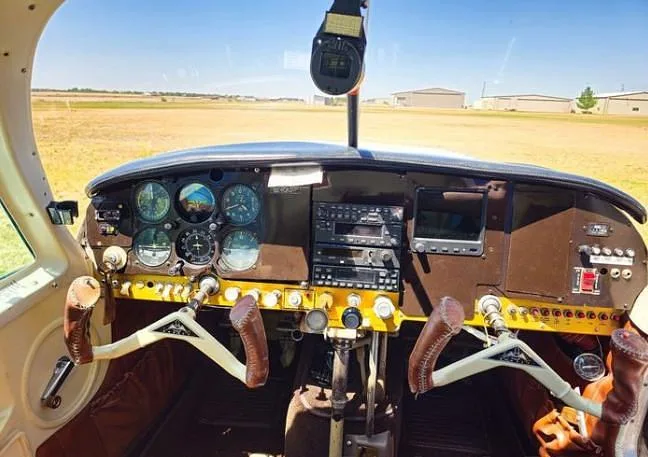 The instrument panel and dual yokes of an aircraft cockpit, with a view of a grassy airfield outside.
