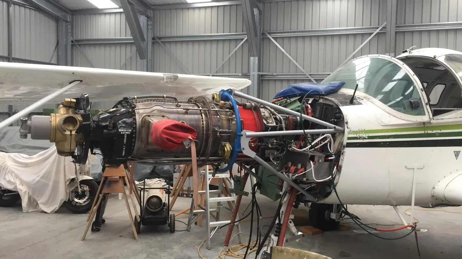 A turboprop aircraft in a hangar with its engine cowling removed, revealing the engine and internal components during maintenance.