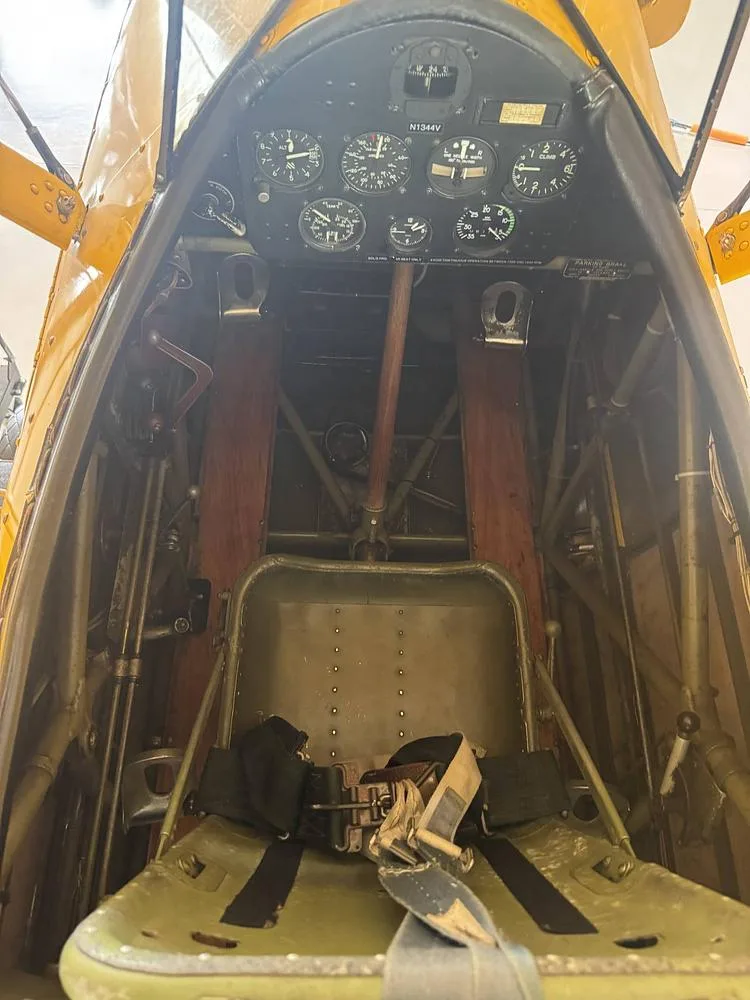 An overhead view of the front cockpit of a yellow Stearman biplane, showing the instrument panel with N1344V and an olive green seat.