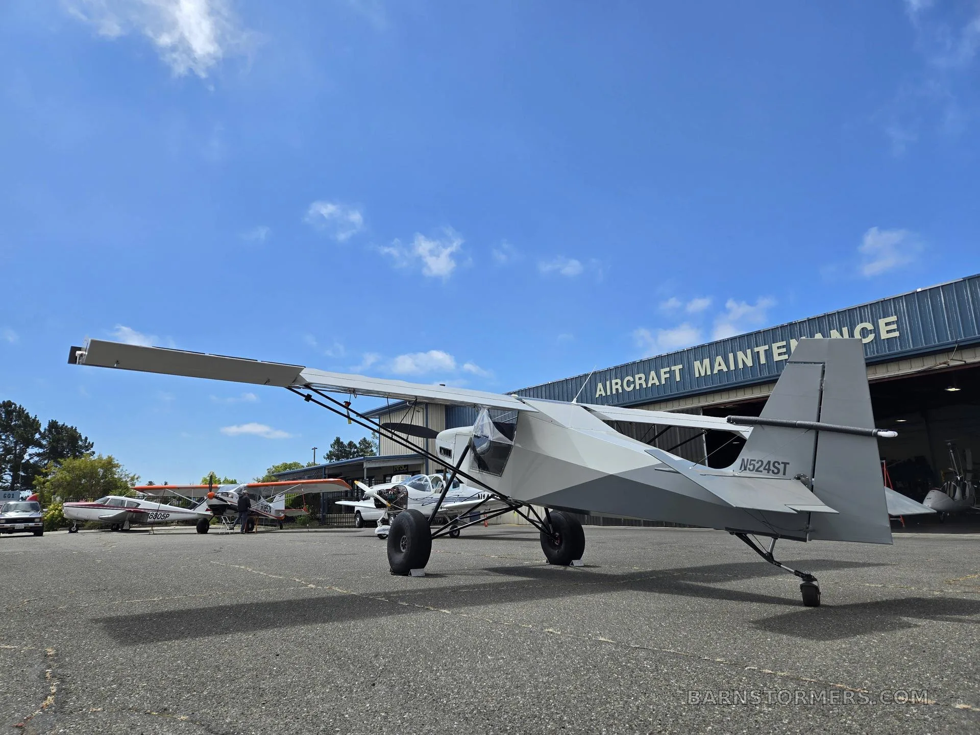 A light gray high-wing aircraft with registration N524ST parked on an airfield in front of an aircraft maintenance hangar.