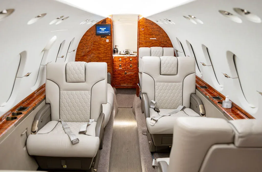 The luxurious interior of a private jet cabin with light grey quilted leather seats and polished wood accents.