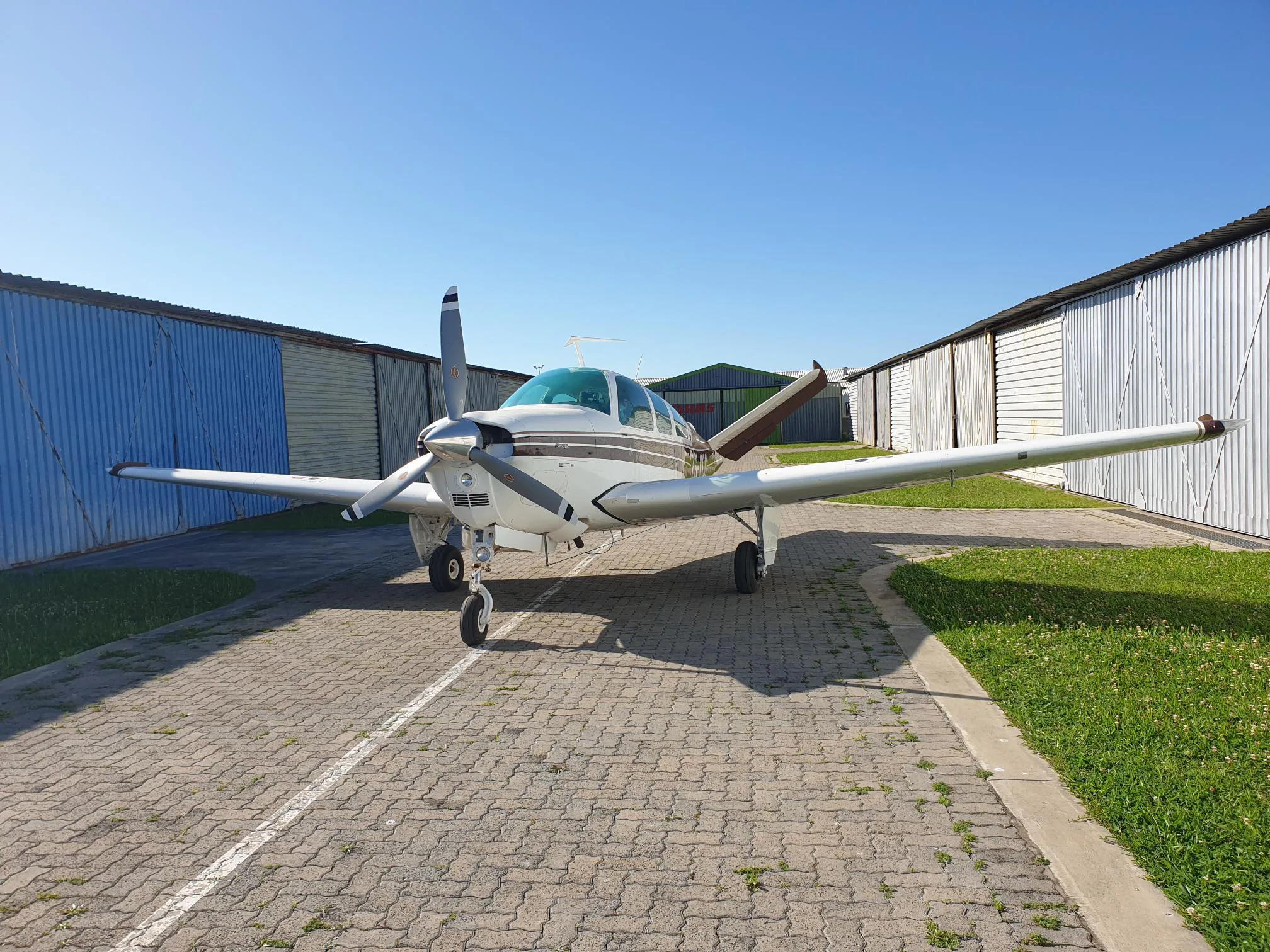 A white and brown single-engine propeller aircraft parked on a paved ramp between two hangars.