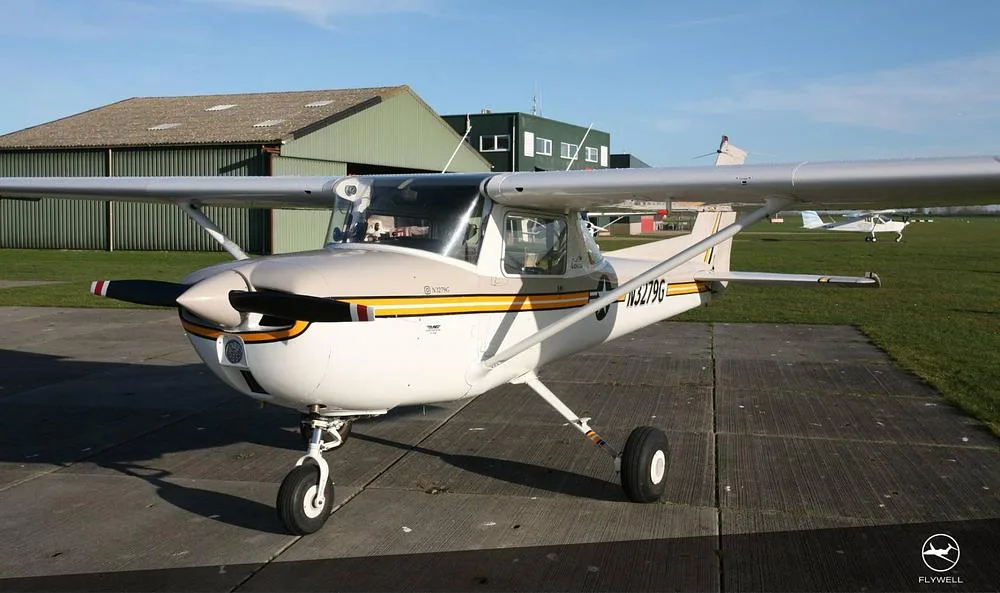 A front-left view of a white and beige Cessna 172 with yellow and black stripes, parked on a concrete pad next to a green hangar.