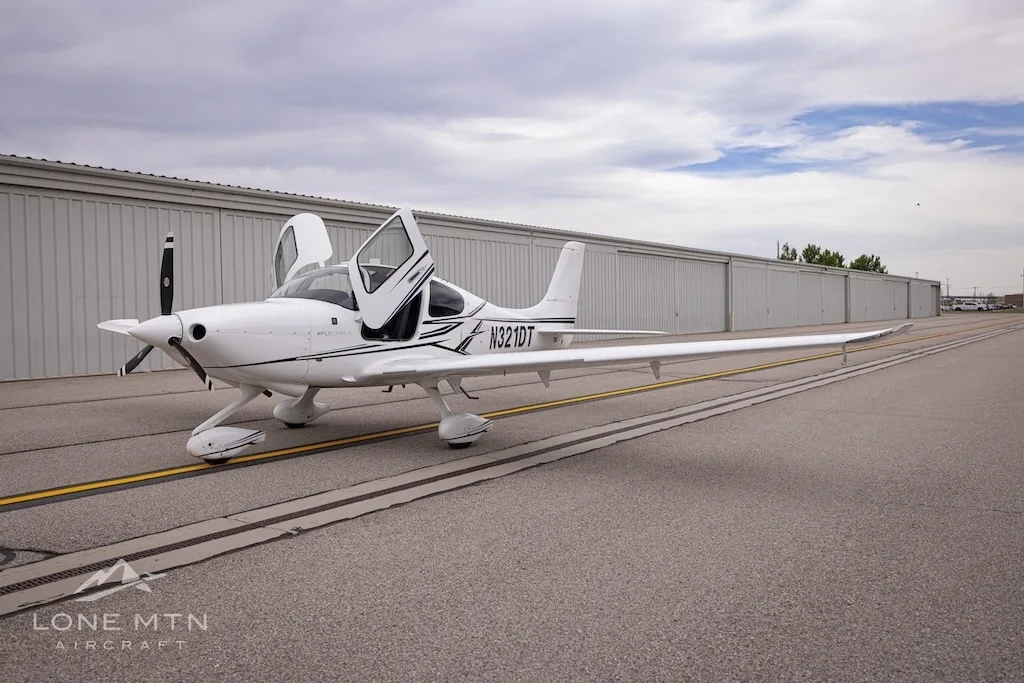 White Cirrus SR22 aircraft with black and grey stripes, doors open, parked on an asphalt ramp in front of hangars.
